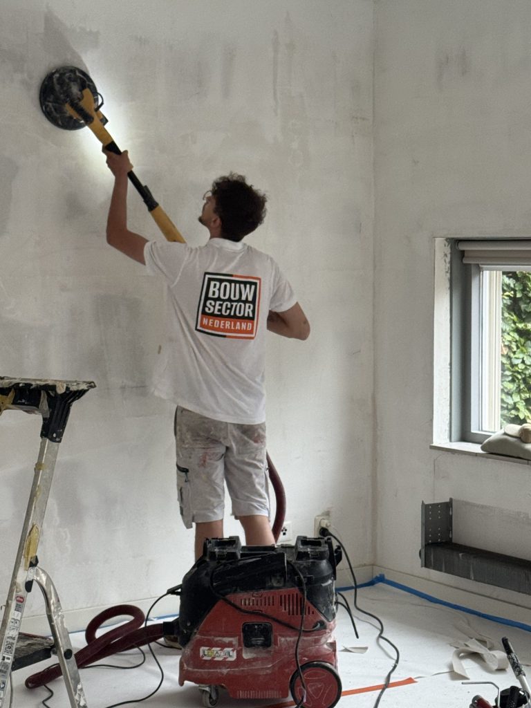 Vakman van Bouwsector Nederland schuurt een wand met een schuurmachine op een steel, de vloer is afgedekt en afgeplakt