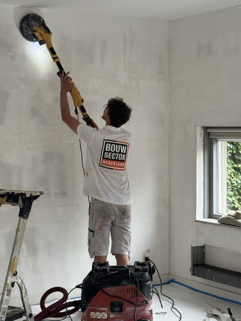 Vakman van Bouwsector Nederland schuurt met een schuurmachine op een steel de bovenkant van een wand, met afzuiging op de afgedekte vloer