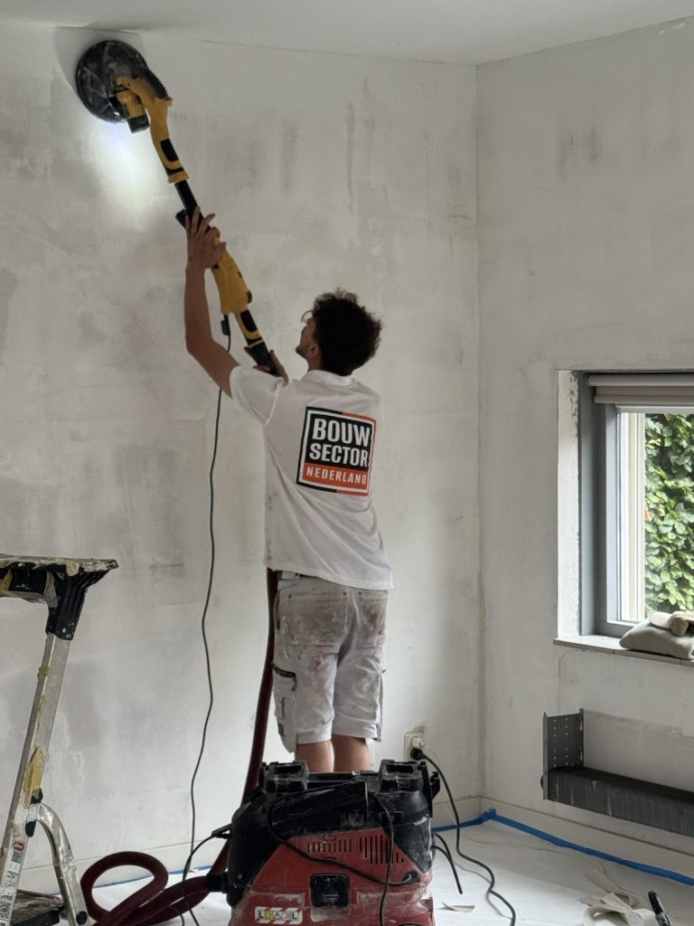 Vakman van Bouwsector Nederland schuurt met een schuurmachine op een steel de bovenkant van een wand, met afzuiging op de vloer