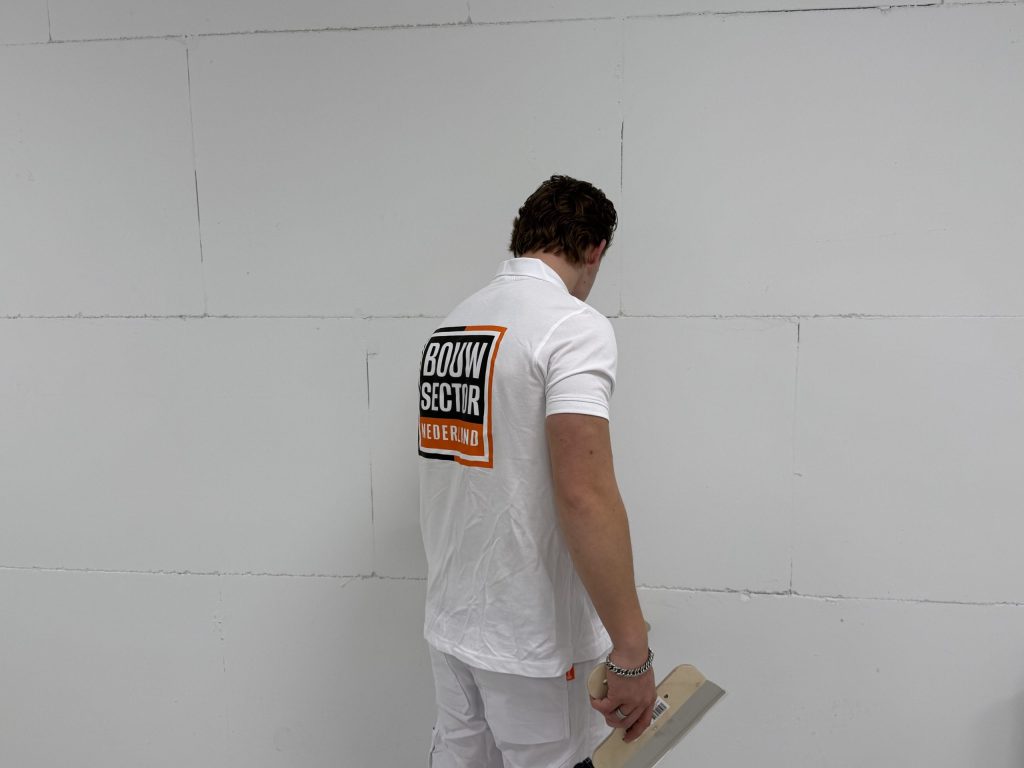 Vakman van Bouwsector Nederland bekijkt met een spaan in de hand een kale wand met zichtbare naden