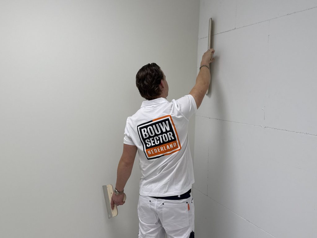 Vakman van Bouwsector Nederland houdt een lange spaan verticaal tegen een wand, met een tweede spaan in zijn andere hand
