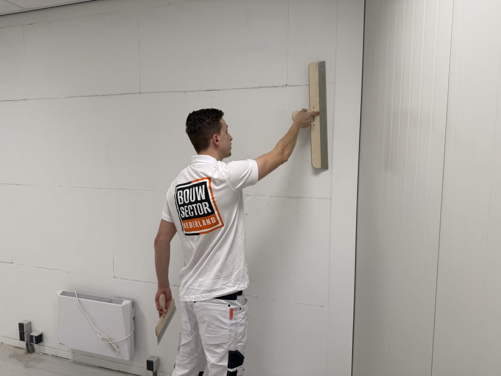 Vakman van Bouwsector Nederland zet een brede spaan hoog tegen een kale wand, met een tweede spaan in zijn andere hand